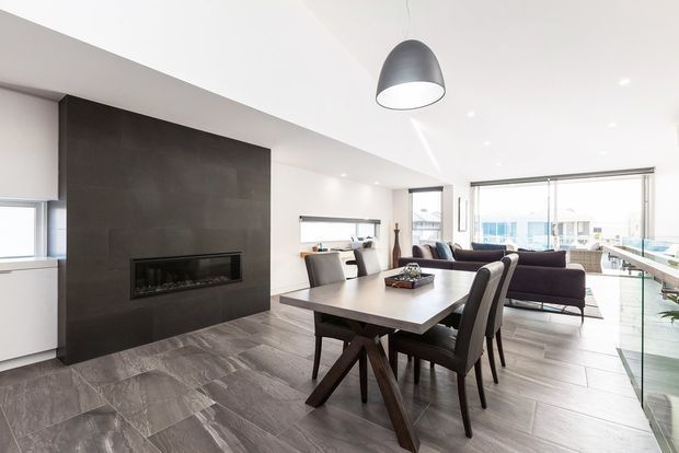 Bright open-plan contemporary apartment interior combining a dining area and living room. A dark feature wall with a linear built-in fireplace anchors the room, while a timber dining table, upholstered chairs, dark sectional seating, large glazed doors and windows, recessed ceiling lights, and a sculptural pendant create a refined modern setting. Grey wood-look flooring runs throughout, with an urban outlook visible through the glazing.