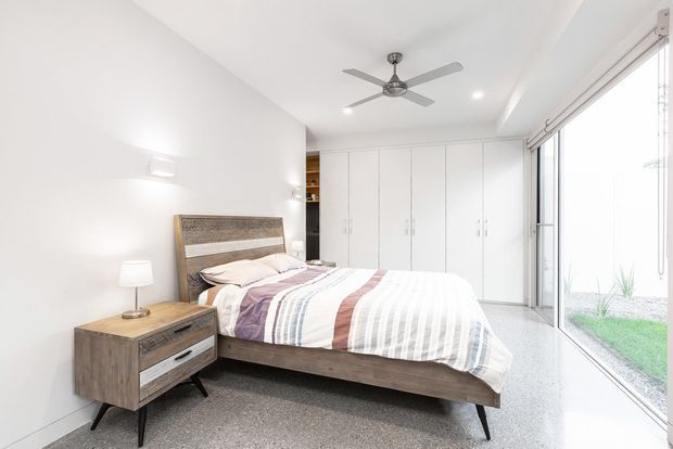 Bright contemporary bedroom with a low timber bed, upholstered striped bedding, matching timber bedside table, full-height white built-in wardrobes, polished concrete-look flooring, recessed lighting, ceiling fan, and extensive floor-to-ceiling glazing overlooking a small planted outdoor area.