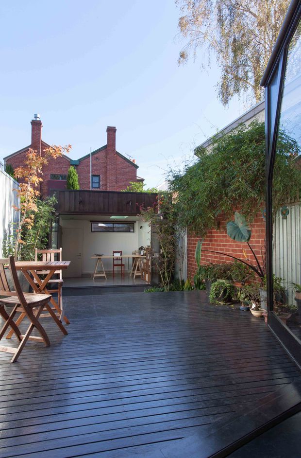 A landscaped urban courtyard viewed from a glazed interior, dominated by a spacious dark timber deck. Folding timber outdoor dining furniture sits to the left, while dense planting, a red brick wall, and a corrugated metal boundary frame the space. At the rear is a small covered outdoor room or studio with a workbench and stools, beneath a dark timber balcony or awning. A neighbouring red-brick pitched-roof house is visible beyond the courtyard.
