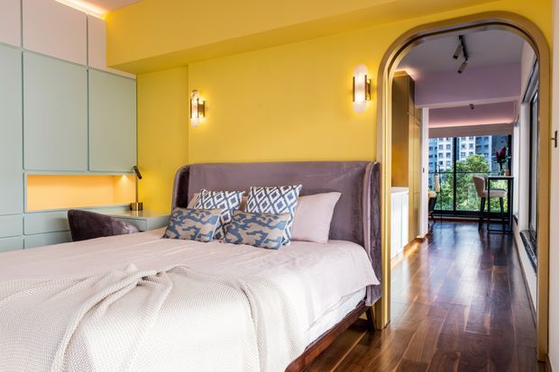 A colourful, compact bedroom with a large upholstered bed, patterned blue-and-white cushions, pale bedding, and warm yellow walls. Built-in pale cabinetry and a small desk area occupy the left side, while an arched doorway leads to a polished timber-floored adjoining space with glazing and additional seating. Warm integrated lighting and brass-toned wall sconces create an inviting, richly styled interior.