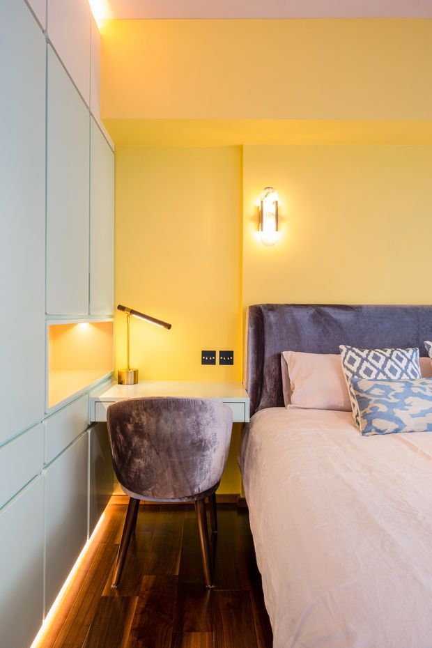 A compact, warmly lit bedroom with a built-in home office nook. The room features full-height pale cabinetry along the left wall, a floating desk with a slim task lamp, a plush upholstered desk chair, a double bed with a tall velvet headboard, patterned cushions, dark timber flooring, and concealed LED strip lighting.