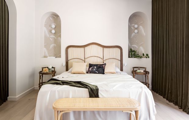 A softly styled bedroom with a central bed framed by a curved natural rattan headboard, white bedding, pale timber flooring, and matching bedside tables. Tall dark curtains frame the room, while two arched wall niches contain subtle decorative relief and lighting. A woven rattan bench sits at the foot of the bed, creating a warm, refined, contemporary interior.