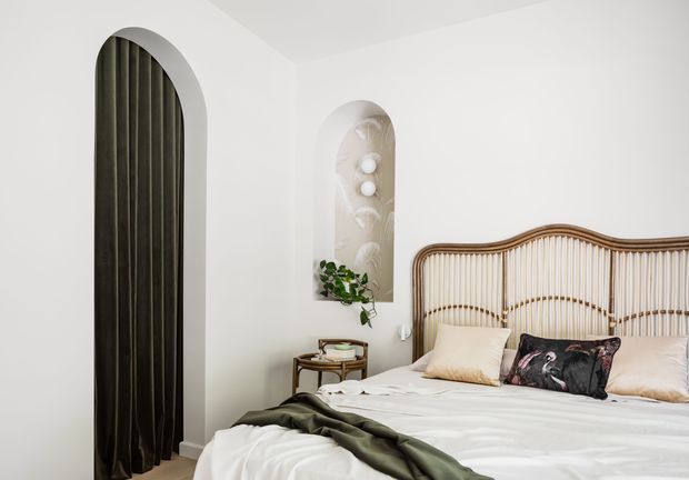 A bright, softly styled bedroom with a large bed positioned against the right wall. The room features a sculptural woven cane/rattan headboard with curved detailing, white bedding, neutral pillows, and a dark olive throw. White walls include two tall arched recessed niches: one conceals dark vertical drapery and the other displays decorative lighting and a small plant. A compact round side table sits beside the bed. The overall look is calm, refined, and organic with strong arched architectural detailing.