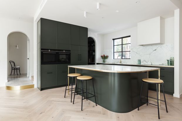 Bright contemporary kitchen with a large dark green built-in cabinetry wall, integrated appliances, and a curved island with a pale stone benchtop and waterfall edge. The island provides casual seating for four on timber-and-black stools. Pale herringbone timber flooring, a white ceiling, marble-look splashback, black-framed window, arched doorway, and subtle brass detailing create a refined, modern interior.