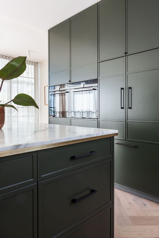 A refined contemporary kitchen with a large dark grey-green built-in cabinetry wall, integrated double ovens, tall refrigerator storage, and a matching island with black bar pulls. The island has a bright polished white marble or engineered-stone benchtop with subtle veining. Pale timber herringbone flooring, sheer curtains, and a large leafy indoor plant add warmth and softness to the otherwise streamlined space.