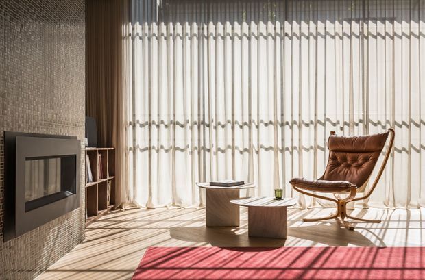 A refined modern living room with a floor-to-ceiling sheer-curtained window, textured tiled feature wall, built-in fireplace, low sculptural coffee tables, open shelving, and a brown leather lounge chair. Strong sunlight creates patterned shadows across the pale floor and red area rug.