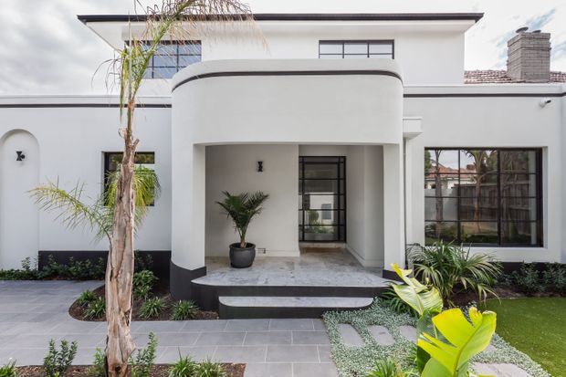 Front exterior of a contemporary white rendered house with a projecting rounded entry canopy, black trim and plinth, large black-framed windows, a tiled roof section, and a landscaped garden with palms and paved paths.