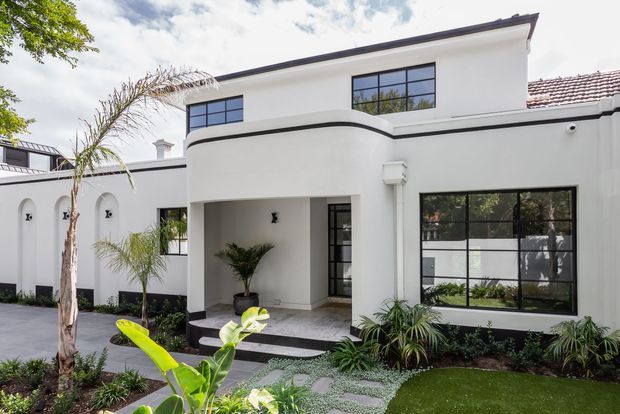 A bright white two-storey residential facade with a curved central entry porch, black-framed grid windows, dark trim, tiled roof sections, and landscaped front garden. The entrance is approached by dark paving, with tropical planting and a manicured lawn in the foreground.