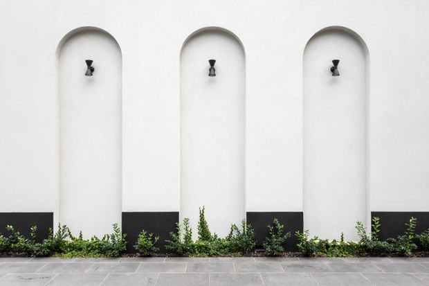 Minimalist architectural exterior featuring a white rendered wall with three tall rounded-arch recessed niches, each containing a small dark wall light. A black lower wall band contrasts with the white upper surface, with low planting along the base and grey rectangular paving in the foreground.
