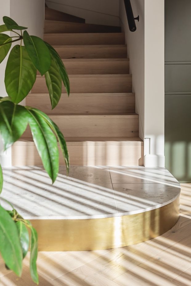 A sunlit interior stairway with broad natural timber stairs rising behind a curved, raised landing or platform. The platform has a pale stone or terrazzo-like top with a polished brass or gold-toned vertical edge, while a dark handrail is mounted along the right wall. Strong linear sunlight and shadows cross the light timber floor and steps, with a large green indoor plant in the foreground and muted built-in cabinetry visible at right.