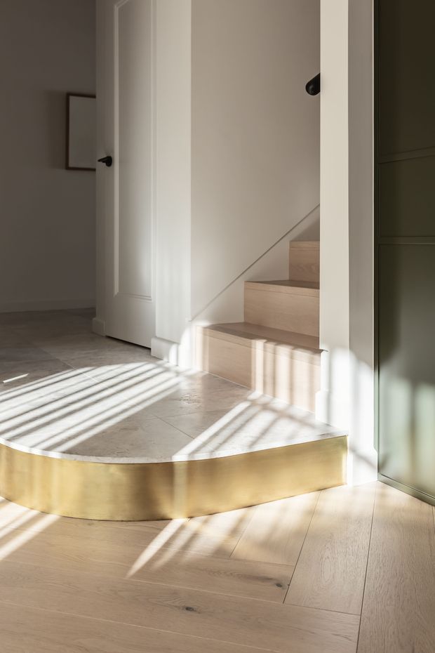 A softly lit interior stair hall with a curved, brass-clad step or threshold transitioning from pale timber flooring to a tiled landing. Pale timber stairs rise beside a white paneled wall, with a white door on the left and dark green built-in cabinetry or wall panelling on the right. Strong sunlight creates striped shadows across the floor and stair surfaces, giving the space a warm, refined atmosphere.
