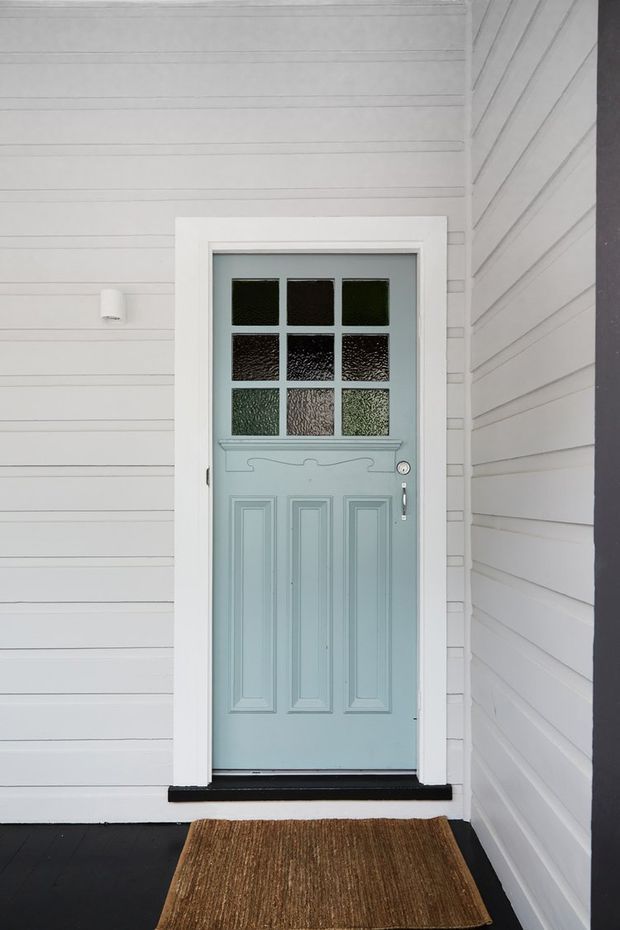 A front entry door set within a white horizontal weatherboard-clad facade. The pale blue-green timber door has decorative recessed panels, a row of nine textured glass panes, white trim, and traditional hardware. A dark porch floor and woven doormat complete the entry.