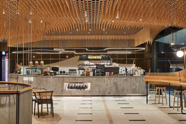 A refined contemporary café or hospitality interior featuring a long service counter, illuminated shelving and signage, extensive warm timber slat detailing across the ceiling and walls, dark glazing, pendant lighting, and a mix of bar stools and café seating. The open floor uses large light tiles with contrasting linear inlays.