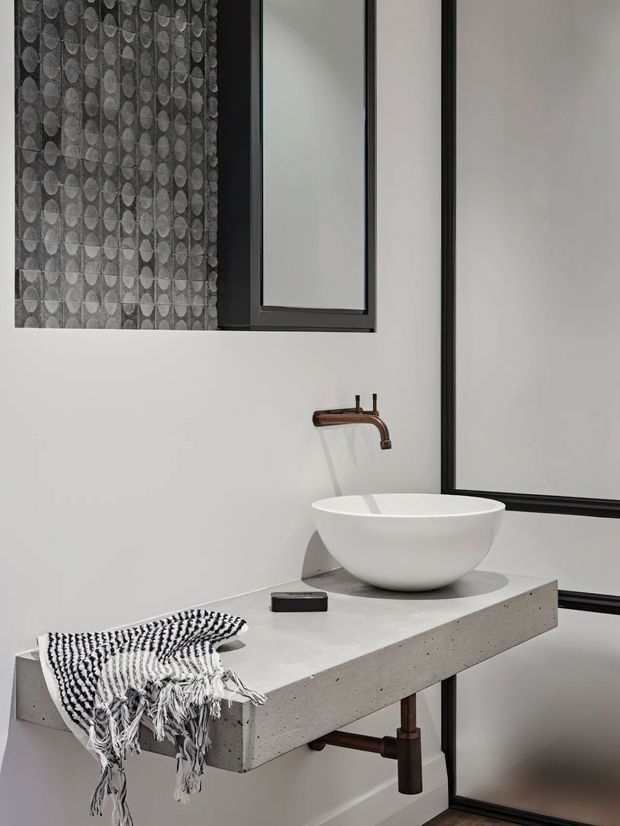 A minimalist contemporary bathroom vanity featuring a white vessel basin on a thick raw concrete benchtop, a wall-mounted copper-toned tap, black-framed mirrors, and a patterned grey feature tile wall. Exposed dark plumbing is visible beneath the floating vanity, with a monochrome tasseled textile draped over the edge.