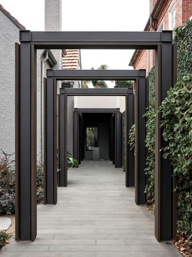 A narrow landscaped outdoor passage framed by a sequence of dark charcoal rectangular portal structures, creating a strong perspective toward a dark entry or courtyard at the far end. Light grey rectangular paving runs through the walkway, bordered by dense hedging and planting. Existing rendered and red-brick building walls with tiled roof sections are visible on either side.