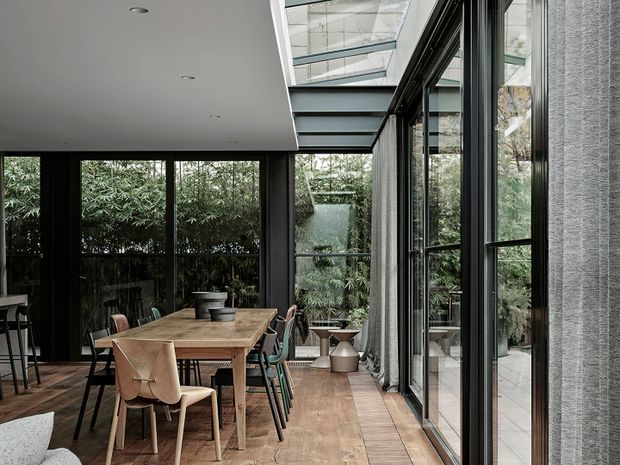 A refined dining area in a contemporary glass-lined room, featuring a long timber dining table with mixed chairs, dark-framed floor-to-ceiling glazing, a large roof skylight, timber flooring, and lush greenery visible outside. The space has a calm, sophisticated indoor-outdoor connection with abundant natural light.