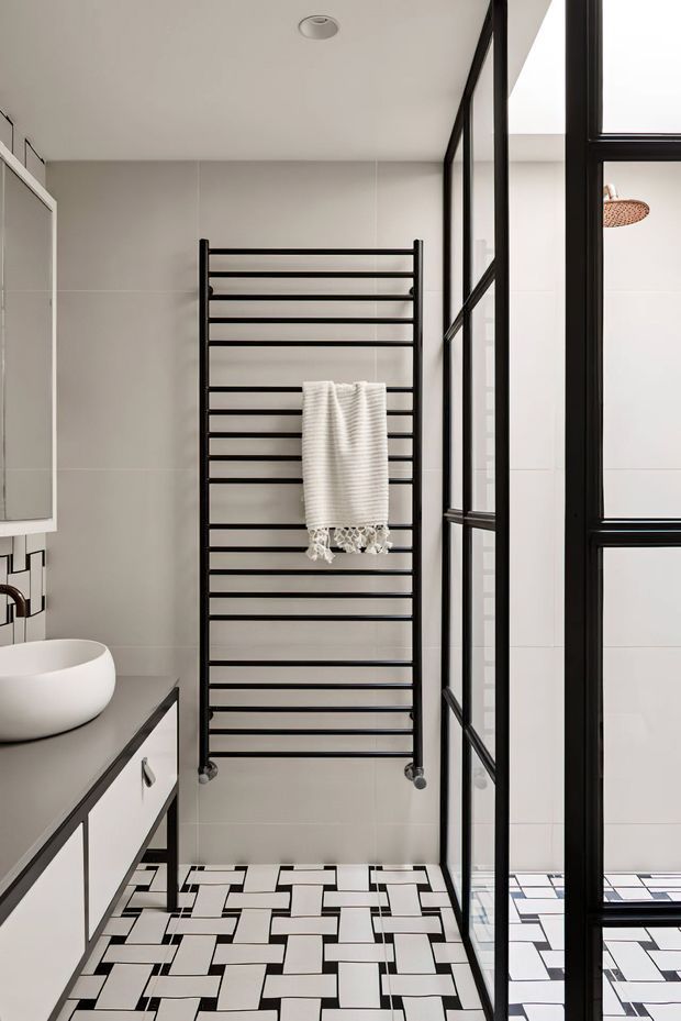 A bright contemporary bathroom featuring a wall-mounted black heated towel rail, white towel, pale grey wall tiles, a floating vanity with a white vessel basin and grey benchtop, and a bold black-and-white geometric tiled floor. Black-framed glass partitions create a shower enclosure or wet area along the right side.