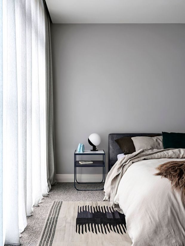 A calm, contemporary bedroom corner with a grey painted wall, floor-to-ceiling sheer curtains, a textured grey carpet, and a partially visible upholstered bed dressed in layered neutral bedding. A slim black metal bedside table holds a spherical lamp and small decorative objects, while a monochrome patterned rug adds graphic contrast.