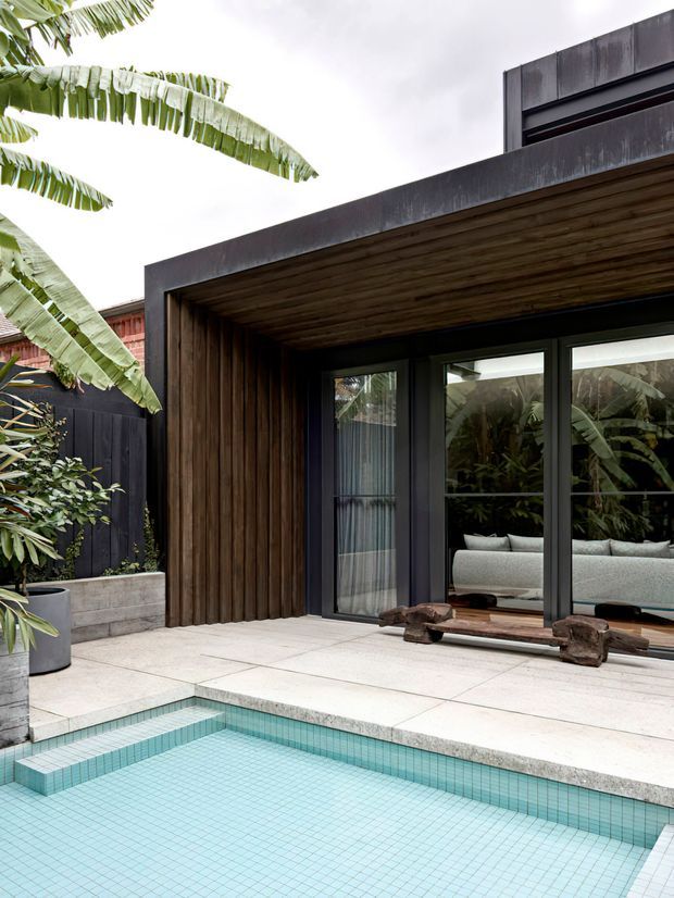 A contemporary rear facade and pool terrace viewed from outdoors. The dark, rectilinear extension has a deep timber-lined roof soffit and matching vertical timber wall cladding, with large black-framed glazed doors opening toward a pale interior. A small square swimming pool with light aqua mosaic tile sits in the foreground, surrounded by pale concrete or stone paving and tropical planting.