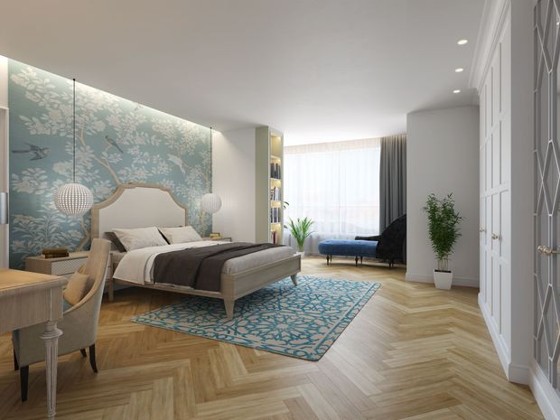 A bright, refined bedroom with a large upholstered bed, patterned blue feature wallpaper, matching blue rug, herringbone timber flooring, a compact desk and chair, built-in shelving, and a small seating area by the window. White built-in wardrobes with decorative glazed doors line the right wall, while sheer curtains provide soft natural light.