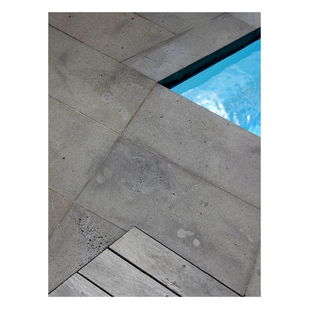 Close-up detail of a contemporary poolside terrace, showing large grey concrete or stone paving slabs with visible aggregate and grout lines, a narrow strip of bright blue swimming pool water, and timber decking at the foreground edge.