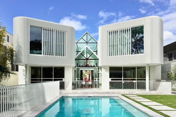 Front exterior view of a contemporary multi-storey residence with two rounded white wings, strong vertical fins, expansive glazing, and a central glazed peaked atrium entry. A rectangular swimming pool, white balustrades, paved stepping stones, and lawn occupy the foreground.