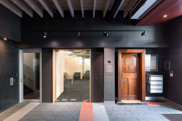 A dark, refined commercial or apartment-building lobby/entry corridor with exposed timber ceiling beams, black walls, track lighting, carpet tiles with a red-orange stripe, and a striking carved timber entry door. Glazed doors reveal a stairwell and a small furnished interior area, while a bank of mailboxes and wall-mounted signage reinforce the shared-entry setting.