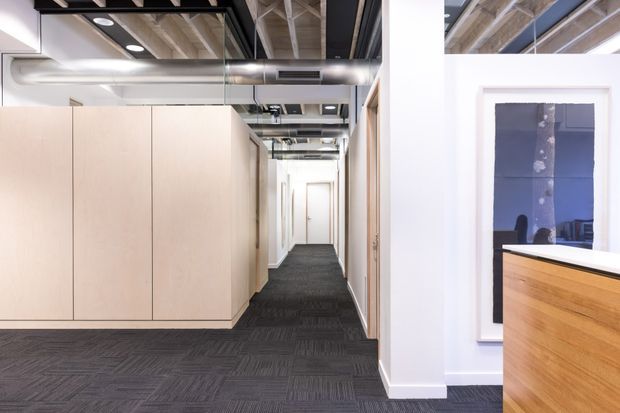 A bright contemporary commercial interior viewed along a long corridor. The space features dark patterned carpet tile flooring, pale timber built-in cabinetry or partition walls, white walls, exposed structural ceiling elements, black beams, recessed lighting, and visible metal HVAC ductwork. A framed artwork and part of a timber reception counter appear at the right.