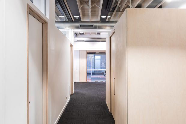 A bright modern commercial hallway or office corridor with pale timber and white partition walls, dark carpet flooring, exposed structural ceiling elements, recessed circular downlights, and visible ductwork. Glazed areas and office spaces appear at the far end. The space has a clean, contemporary workplace fit-out with contrasting dark flooring and light joinery.