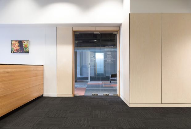 A contemporary interior hallway or entry space with dark patterned carpet flooring, pale neutral walls, and extensive light timber built-in cabinetry. A large timber-fronted console or reception unit sits to the left, while a framed glazed doorway opens toward another modern interior beyond. The space is clean-lined and restrained, with subtle artwork on the wall and concealed storage.
