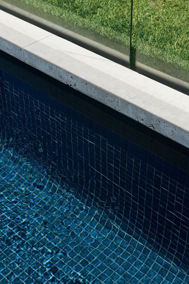 Close-up detail of a swimming pool with deep blue square mosaic tile lining, visible rippling water, and a pale textured stone or concrete coping edge beside a frameless glass barrier. Green lawn is visible beyond the glass.