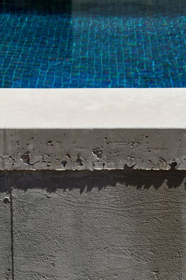 Close-up architectural detail of a swimming pool edge, showing blue tiled pool water above a pale coping surface and a rough, exposed concrete pool wall below. The image emphasizes the contrast between smooth light coping, textured concrete, and deep blue water.