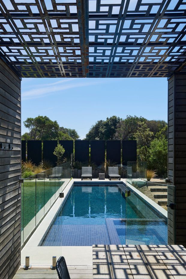 A modern outdoor pool courtyard viewed from a timber-clad structure, with a narrow blue-tiled swimming pool, adjacent shallow water feature, frameless glass fencing, timber decking, dark privacy screens, sculptural planting, and a striking geometric overhead pergola casting patterned shadows.