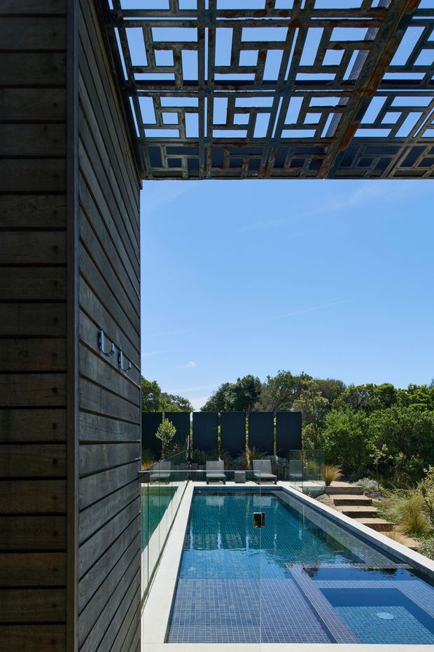 A contemporary outdoor swimming pool viewed from beside a dark horizontal timber-clad wall. The pool is lined with small blue tiles and bordered by pale paving, with glass balustrading, lounge seating, landscaped planting and a striking overhead timber pergola featuring an open geometric lattice pattern.