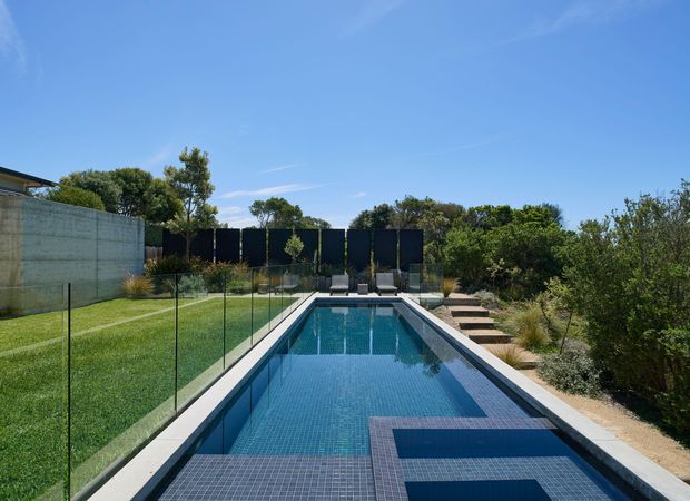 A long, narrow rectangular swimming pool with dark blue tiled surfaces and geometric contrasting in-pool detailing, bordered by pale concrete coping. Frameless glass pool fencing runs along the manicured lawn on the left, while a landscaped garden with stepping stones and dense shrubs borders the right. Dark privacy screening and outdoor lounge chairs sit at the far end beneath a clear blue sky.