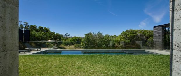 A contemporary landscaped courtyard viewed between two concrete walls, with a long rectangular swimming pool, expansive lawn, dense greenery beyond, and a dark-clad pavilion or pool structure at the right. The composition emphasizes the horizontal pool, open sky, and strong concrete framing.