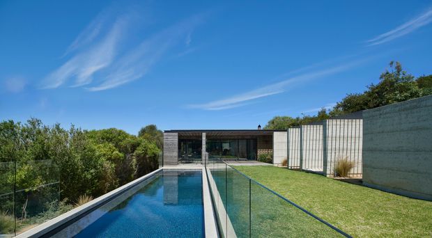 Exterior view of a contemporary single-storey residence set among dense greenery, with a long narrow swimming pool, transparent pool safety fencing, lawn, and a low flat-roofed building in the background. The home features dark framed glazing and pale textured concrete walls under a clear blue sky.