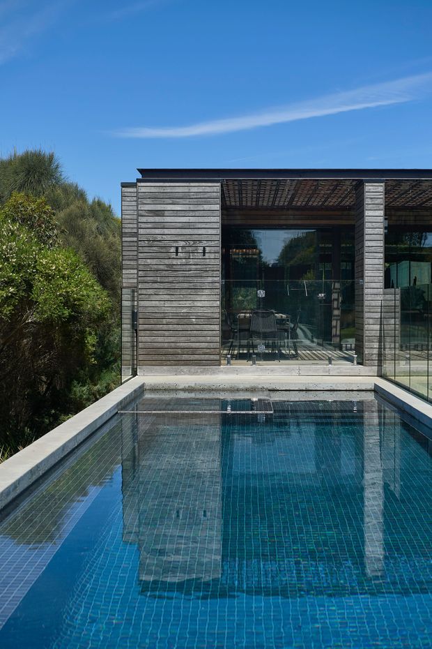 Exterior architectural view of a modern, dark timber-clad pavilion beside a rectangular swimming pool. The building features extensive floor-to-ceiling glazing, a flat roof with a covered outdoor area, exposed timber screening or rafters, and a concrete pool surround. Dense coastal-style vegetation borders the left side.
