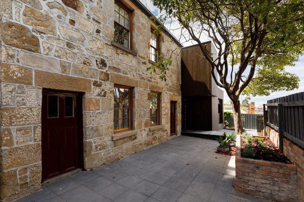 A sunlit courtyard beside a two-storey stone building. The facade is constructed from irregular pale natural stone blocks with contrasting mortar, timber-framed windows, and a dark timber entry door. A contemporary dark vertical-clad extension or structure meets the historic stonework, while large grey paving slabs, brick-edged raised planting beds, and mature trees define the courtyard.