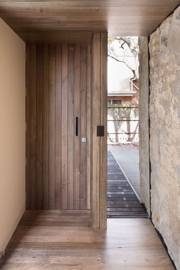 A refined entryway with a tall, vertically timber-panelled pivot door and matching timber-lined ceiling and floor. A rugged exposed stone wall forms the right side of the passage, opening toward a small exterior courtyard with timber decking. The restrained material palette creates a warm, tactile, contemporary-rustic character.