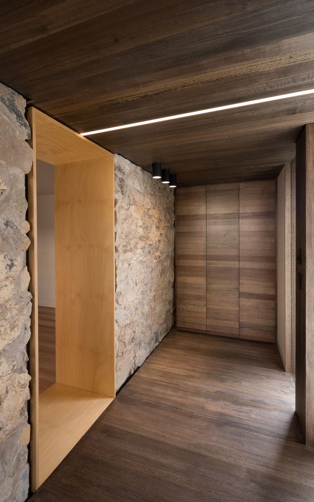 A warm, narrow interior circulation space with dark timber flooring and a matching timber-panelled ceiling. A rough exposed stone wall runs along the left side, contrasted by a light timber built-in alcove or doorway surround. At the end of the passage is a full-height bank of dark wood storage cabinetry with subtle vertical divisions. Linear recessed lighting and small black downlights illuminate the corridor, creating a refined rustic-modern atmosphere.