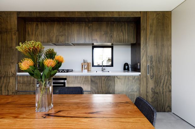 A warm, contemporary open-plan kitchen and dining area. Dark timber cabinetry and wall panelling frame a compact kitchen with a pale benchtop, integrated cooktop, sink, small window, and black appliances. In the foreground, a large natural timber dining table with woven dark chairs and a vase of flowers creates a strong dining zone.