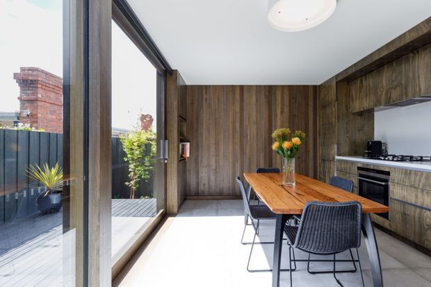 A contemporary dining and kitchen interior with a long timber dining table, dark woven chairs, full-height timber wall panelling, and matching timber kitchen cabinetry. Large glazed doors open onto a small deck or courtyard, bringing in abundant daylight. The space includes a compact kitchen with integrated appliances, a pale smooth floor, and a circular flush-mount ceiling light.