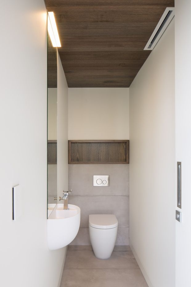 Compact contemporary powder room with a wall-hung white basin and toilet, large-format grey concrete-look wall and floor tiles, a dark timber ceiling, and a recessed timber storage panel above the concealed cistern. A full-height mirror and slim linear wall lighting contribute to the clean, minimalist fit-out.