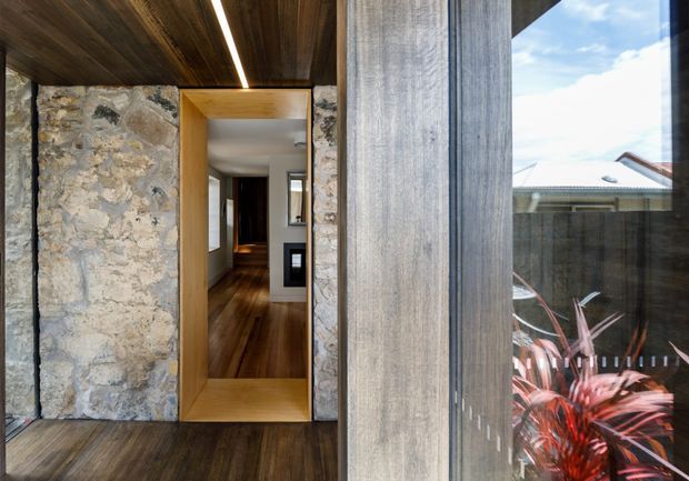 A contemporary residential entry viewed from outside or just within the threshold, with a large timber-framed doorway set into rough natural stone walls. Dark timber ceiling and flooring continue into a narrow hallway, while a tall glazed opening reveals a courtyard planting area and neighbouring roof beyond. The composition highlights tactile stone, warm timber, strong vertical framing, and integrated linear lighting.