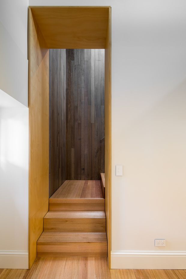 A narrow, built-in timber stairway rises through a rectangular opening in a pale plaster wall. The stairs, side panels, and overhead soffit are finished in warm honey-toned wood, while the upper stair enclosure features a darker vertical timber lining. White skirting boards, a light switch, and a power outlet are visible on the surrounding wall. The image focuses on a compact, highly integrated interior stair detail.