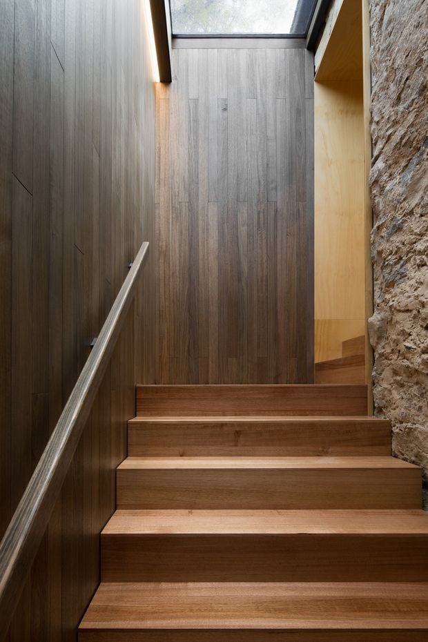 A narrow, contemporary stairway ascending through a vertically lined timber-clad enclosure. The stairs and handrail are warm medium-toned timber, with dark timber wall panelling on the left, a rough exposed stone wall on the right, and a skylight above bringing in natural light. A timber-lined landing or opening is visible at the top.