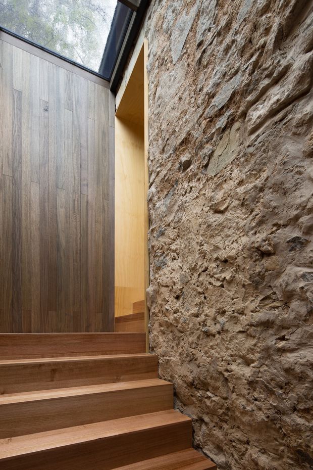 Interior view of a compact stairway with warm timber steps and matching vertical timber wall panelling. A highly textured exposed natural stone wall runs alongside the stairs, while a large sloping skylight or glazed roof panel brings in diffuse daylight. The composition highlights the contrast between smooth timber joinery and rugged masonry.