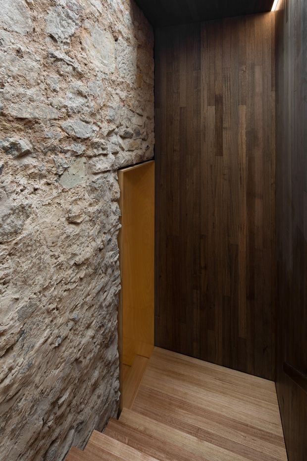 A narrow, moody stairway descending between a rough exposed-stone wall and dark timber-clad walls. The stair treads and landing are finished in light timber, while concealed linear lighting highlights the upper edge of the wood panelled wall. A small timber door or opening is visible at the lower landing.