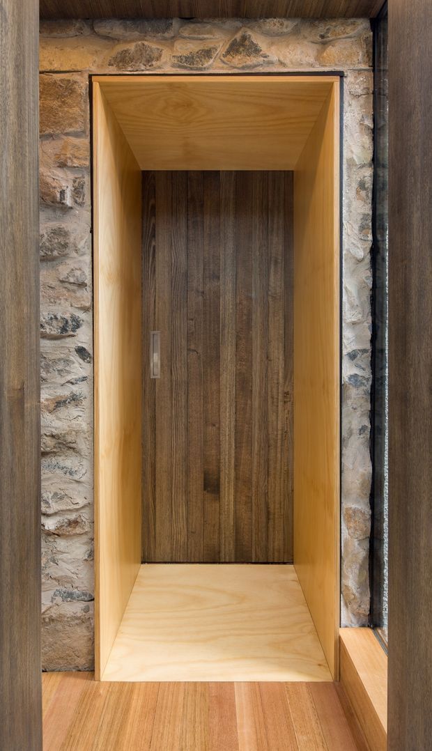 Close-up architectural photograph of a dark timber pivot or entry door recessed within a light timber-lined surround. Rough natural stone masonry frames the opening, while dark timber wall cladding and timber flooring reinforce the warm, tactile material palette. The composition focuses on the built-in doorway and contrasting wood and stone finishes.